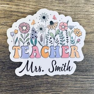 May include: A white sticker with a colorful floral design and the word "TEACHER" in rainbow colors. The text "Mrs. Smith" is written below the word "TEACHER".