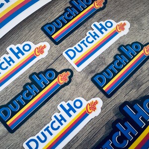 May include: A collection of stickers with the text "Dutch Ho Coffee" in blue, yellow, and red. The stickers have a retro design with a blue border and are arranged on a wooden surface.