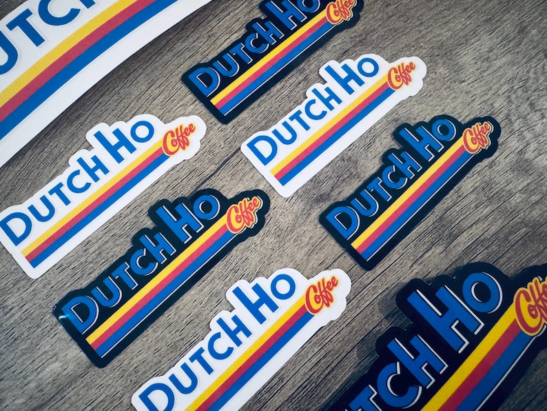 May include: A collection of stickers with the text "Dutch Ho Coffee" in blue, yellow, and red. The stickers have a retro design with a blue border and are arranged on a wooden surface.