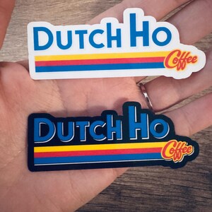 May include: Two stickers with the text "Dutch Ho Coffee". The stickers have a rectangular shape with a blue border and the words "Dutch Ho" in blue. Below the text are three horizontal stripes in red, yellow, and blue. The word "Coffee" is in a stylized font.
