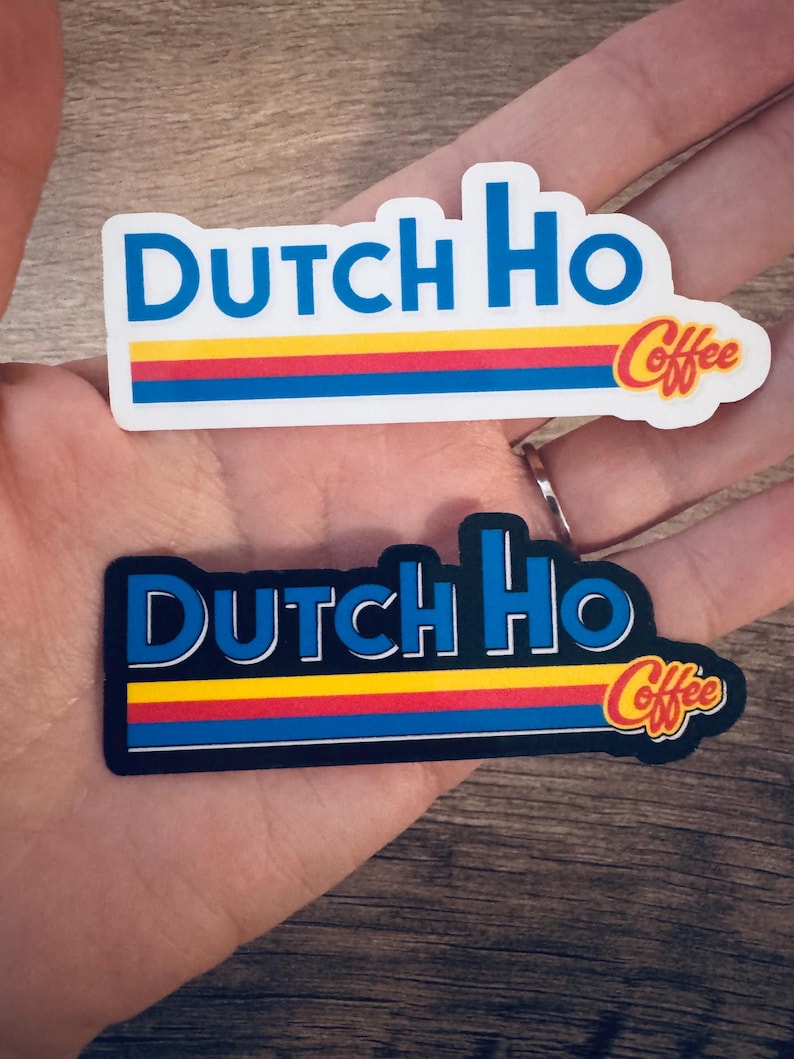 May include: Two stickers with the text "Dutch Ho Coffee". The stickers have a rectangular shape with a blue border and the words "Dutch Ho" in blue. Below the text are three horizontal stripes in red, yellow, and blue. The word "Coffee" is in a stylized font.