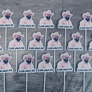 Puede incluir: Un conjunto de catorce adornos para cupcakes id&eacute;nticos, cada uno con un recorte de un hombre con sombrero de vaquero y camisa beige, con el texto "Look who's 44" impreso debajo. Cada adorno est&aacute; sujeto a un palito blanco para su exhibici&oacute;n.