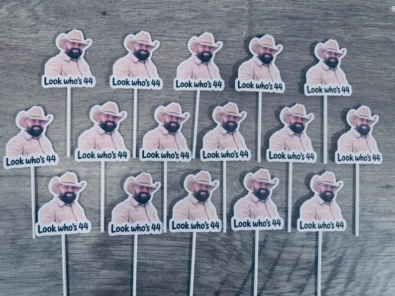 Puede incluir: Un conjunto de catorce adornos para cupcakes id&eacute;nticos, cada uno con un recorte de un hombre con sombrero de vaquero y camisa beige, con el texto "Look who's 44" impreso debajo. Cada adorno est&aacute; sujeto a un palito blanco para su exhibici&oacute;n.