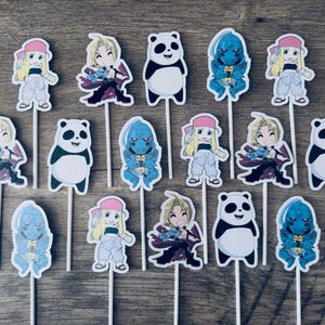 Puede incluir: Un conjunto de quince adornos para cupcakes con ilustraciones de personajes rubios, figuras con armadura azul y pandas en blanco y negro. Cada recorte est&aacute; unido a un palillo blanco fino, mostrando varios dise&ntilde;os de estilo anime.