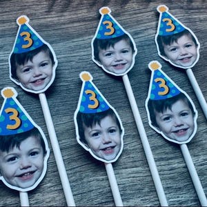 Puede incluir: Cinco adornos personalizados para pasteles con la fotograf&iacute;a del rostro de un ni&ntilde;o peque&ntilde;o con un gorro de fiesta azul de lunares verdes y el n&uacute;mero 3 en amarillo. Cada recorte est&aacute; unido a un palito blanco para colocarlo.