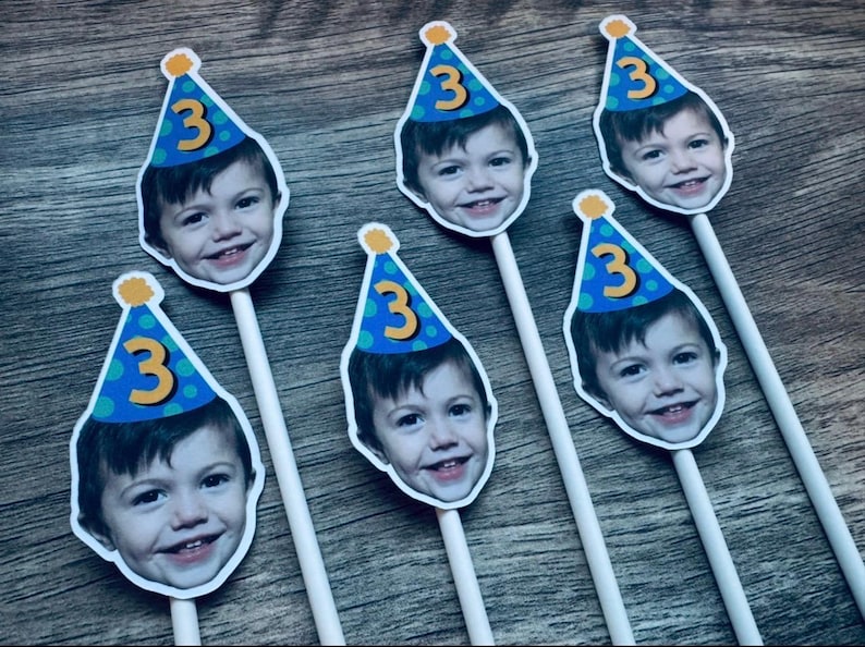 Puede incluir: Cinco adornos personalizados para pasteles con la fotograf&iacute;a del rostro de un ni&ntilde;o peque&ntilde;o con un gorro de fiesta azul de lunares verdes y el n&uacute;mero 3 en amarillo. Cada recorte est&aacute; unido a un palito blanco para colocarlo.