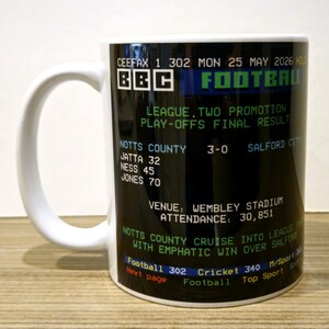 May include: A white ceramic mug featuring a black graphic print that mimics a vintage BBC Ceefax teletext screen. The text displays a football match result between Notts County and Salford City, including the date Monday 25 May 2026 and match stats.