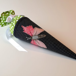 Könnte beinhalten: Eine schwarze und weiße Tupfen-Kegelform aus Stoff mit einem grünen und weißen Tupfen-Oberteil. Ein rosa Schmetterling mit grauem Körper ist auf der Vorderseite der Tasche gestickt.