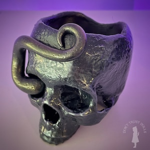 Puede incluir: Un macetero de calavera negro con un diseño de serpiente dorada. El macetero está hecho de un material texturizado y tiene una pequeña abertura en la parte superior para plantar.