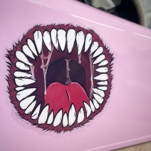 May include: A pink sticker with a cartoon image of a large, open mouth with sharp teeth and a red tongue. The mouth is dripping with saliva.