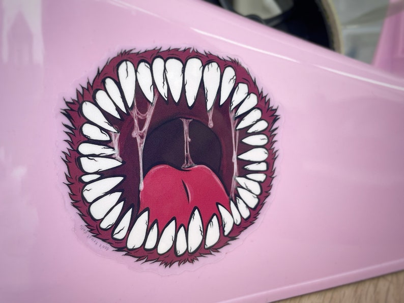 May include: A pink sticker with a cartoon image of a large, open mouth with sharp teeth and a red tongue. The mouth is dripping with saliva.