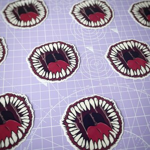 May include: A set of stickers featuring a cartoon mouth with sharp teeth and a red tongue. The stickers are printed on white paper and have a glossy finish.