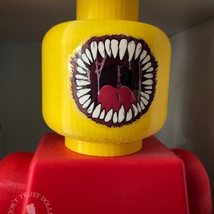 May include: A yellow Lego figure with a red torso. The figure's head has a sticker of a large, open mouth with sharp teeth and a red tongue.