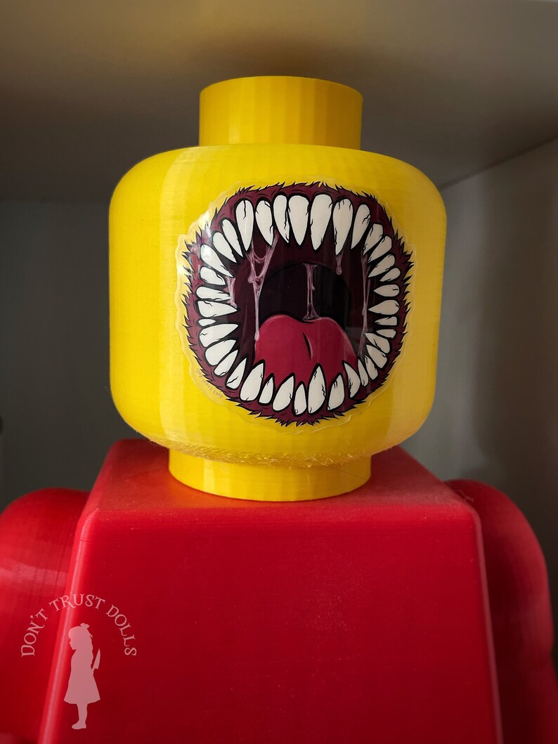 May include: A yellow Lego figure with a red torso. The figure's head has a sticker of a large, open mouth with sharp teeth and a red tongue.