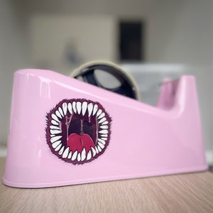 May include: A pink tape dispenser with a cartoon sticker of a mouth with sharp teeth and a red tongue.