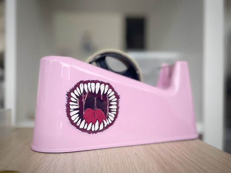 May include: A pink tape dispenser with a cartoon sticker of a mouth with sharp teeth and a red tongue.