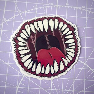 May include: A sticker of a large, open mouth with sharp, white teeth and a red tongue. The mouth is surrounded by a red, furry border.