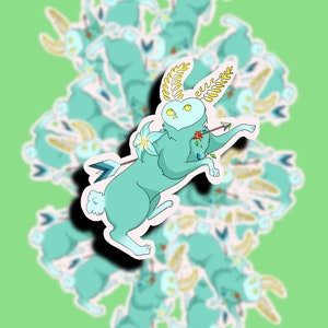 May include: A sticker featuring a light blue rabbit with a flower and an arrow through its body. The rabbit has golden leaves on its head and a heart-shaped face with large yellow eyes. The sticker is surrounded by multiple copies of itself, all slightly out of focus.