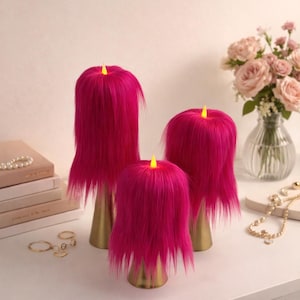 May include: Three decorative candles with bright pink faux fur and a gold base. The candles have a yellow LED light at the top, resembling a flame. The candles are arranged on a white surface with other decorative items.