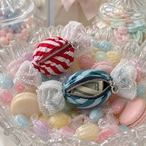 May include: Two candy-shaped zippered pouches with white lace ends. One pouch features red and white stripes, while the other is teal and white with a visible paper bill inside. Both are nestled among assorted pastel-colored candies in a glass bowl.