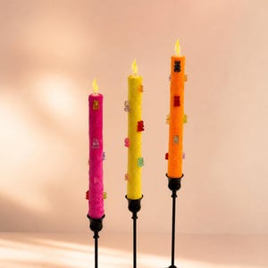 May include: Three colorful decorative candles in pink, yellow, and orange, each adorned with gummy bear accents. The candles are mounted on black metal candlestick holders, creating a playful and vibrant display. The flames are illuminated.