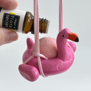 Peut inclure: Un diffuseur d'air en forme de flamant rose avec un cordon rose, une boule rose et une bouteille marron avec le texte "flora secret". Le flamant rose a un bec orange et des yeux noirs. Le flacon est versé dans le flamant.