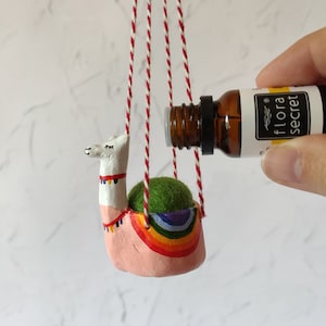 May include: A handmade llama planter in pink, white, and rainbow hues, suspended by red and white twine. The planter contains a green, spherical object. A brown bottle with the text "flora secret" is held near the planter.