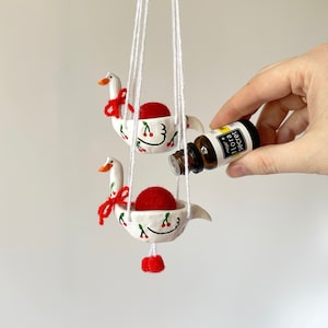 May include: Two white ceramic goose-shaped diffusers hang from white strings. Each diffuser has a red felt ball inside and is decorated with red cherries. A small bottle of essential oil is being poured into one of the diffusers.