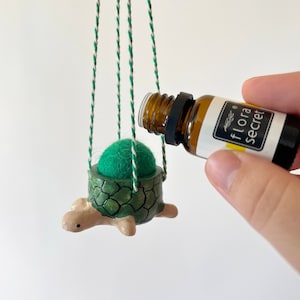 May include: A green ceramic turtle with a felt ball inside is hanging from a string. A hand is holding a small brown bottle with a black label that says "flora secret".