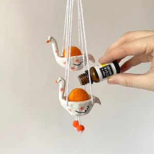 May include: White ceramic swan-shaped diffuser with orange felt balls hanging from a white string. A hand is holding a small bottle of essential oil labelled "flora secret" and pouring it into the diffuser.