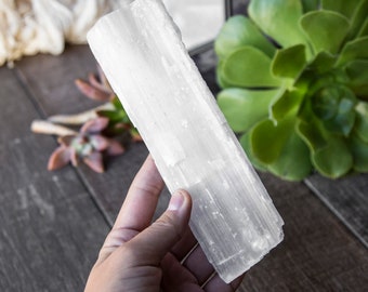 May include: A white selenite crystal wand with a smooth, striated surface. The wand is held in a hand against a wooden background.