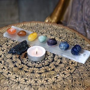 May include: A set of seven chakra crystals arranged on a selenite charging plate with a lit candle in the center. The crystals are red, orange, yellow, green, blue, indigo, and violet. The plate is on a wooden surface with a black and white mandala design.