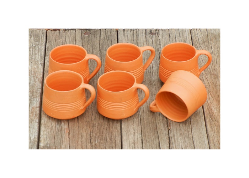 May include: Six terracotta mugs with a ribbed design. The mugs are arranged in a semi-circle on a wooden surface.