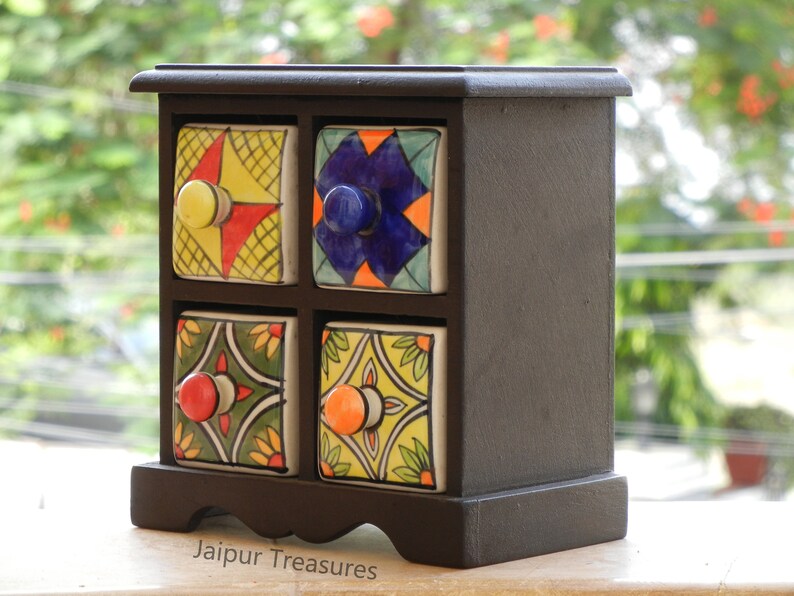 May include: A small wooden box with six colorful ceramic drawers. Each drawer has a different pattern and a knob. The box is on a wooden surface. Jaipur Treasures.