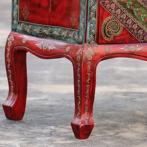 May include: A red wooden cabinet with intricate gold and green painted designs. The cabinet has four legs with ornate details and a drawer with a similar design.