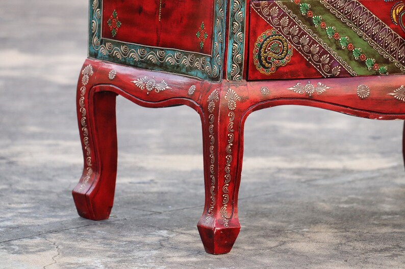 May include: A red wooden cabinet with intricate gold and green painted designs. The cabinet has four legs with ornate details and a drawer with a similar design.