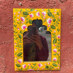 Puede incluir: Un espejo de madera amarillo con un dise&ntilde;o floral pintado a mano con flores rosas y hojas verdes. El espejo tiene un arco decorativo en la parte superior.