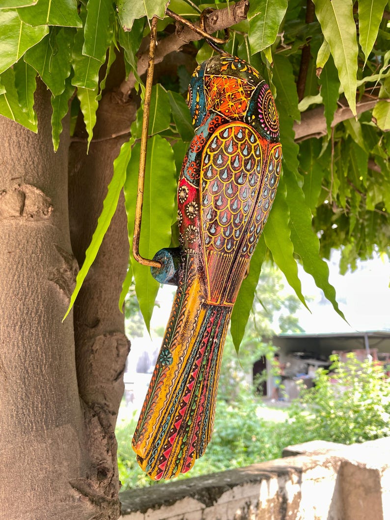 May include: A colorful wooden bird ornament with intricate details and a hanging loop. The bird is painted in shades of yellow, orange, red, blue, green, and black.