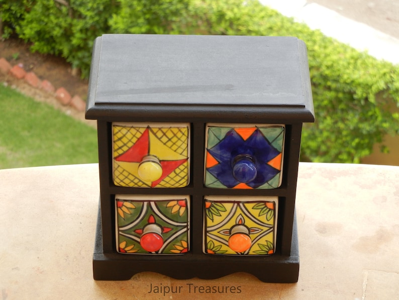 May include: A black wooden box with four small drawers. Each drawer has a colorful ceramic front with a different geometric pattern and a knob. The box is labeled "Jaipur Treasures".