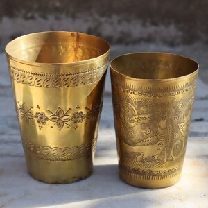May include: Two gold metal drinking glasses with intricate engraved designs. The glass on the left has a floral pattern, while the glass on the right has a more abstract design.