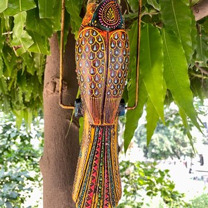 May include: A colorful wooden bird ornament with intricate details, painted in shades of yellow, red, blue, and green. The bird is hanging from a metal ring and has a long tail with a geometric pattern.