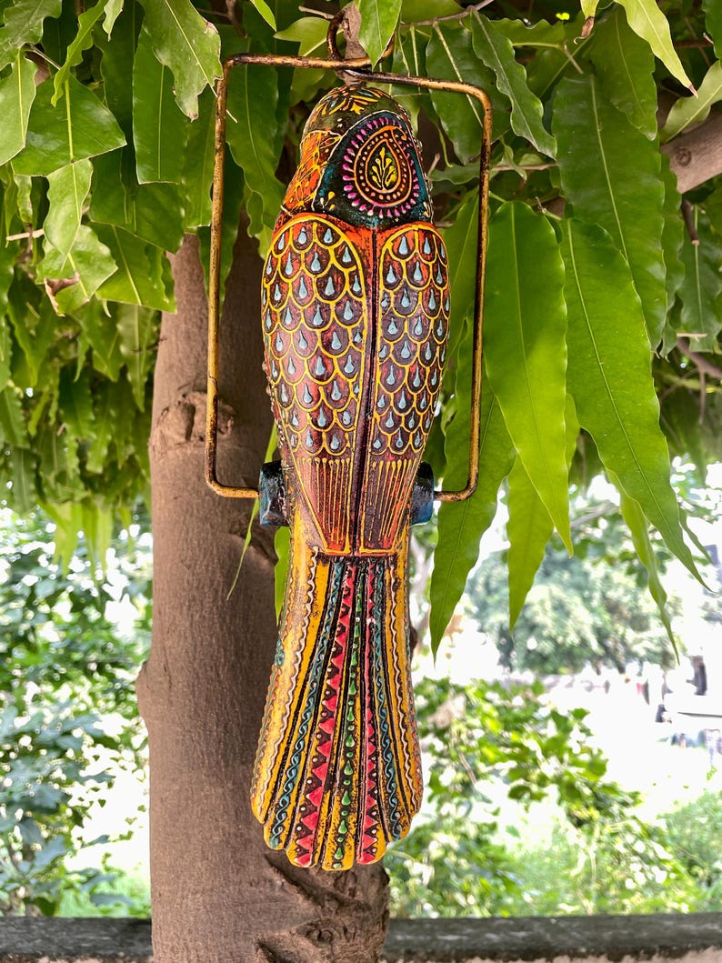 May include: A colorful wooden bird ornament with intricate details, painted in shades of yellow, red, blue, and green. The bird is hanging from a metal ring and has a long tail with a geometric pattern.