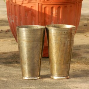 May include: Two antique brass cups with a hammered finish and a dotted pattern. The cups are sitting on a concrete surface.