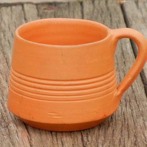 May include: A terracotta mug with a rounded handle. The mug has a textured surface with horizontal lines.