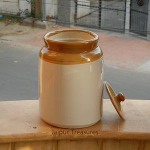 May include: A white ceramic jar with a brown lid. The jar has a brown band around the top. The jar is sitting on a wooden surface. The text "Jaipur Treasures" is printed on the surface.