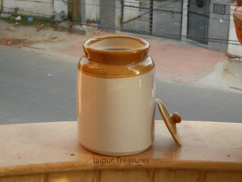 May include: A white ceramic jar with a brown lid. The jar has a brown band around the top. The jar is sitting on a wooden surface. The text "Jaipur Treasures" is printed on the surface.