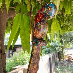 May include: A colorful wooden parrot ornament with intricate details and a hanging loop. The parrot is painted in shades of blue, green, red, yellow, and orange, with a paisley pattern on its body.