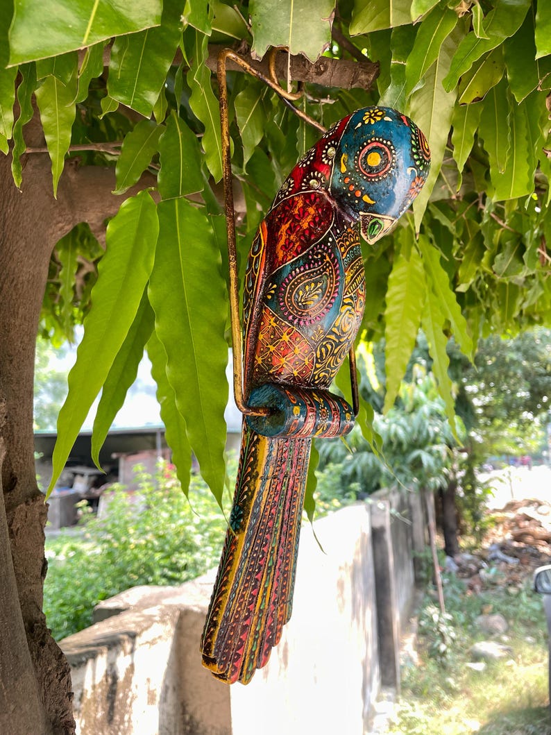 May include: A colorful wooden parrot ornament with intricate details and a hanging loop. The parrot is painted in shades of blue, green, red, yellow, and orange, with a paisley pattern on its body.