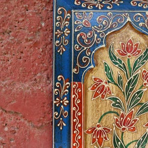 以下が含まれることがあります： 花柄のデザインが施された木製パネルのクローズアップ。パネルは、複雑な模様の青い縁取り、赤と金の中心部、緑の葉を持つ赤い花が特徴です。背景はテクスチャ加工された赤色です。