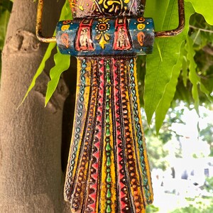 May include: A colorful wooden bird ornament with intricate details and a hanging loop. The bird is painted in shades of blue, yellow, red, green, and brown, with a variety of patterns and designs.